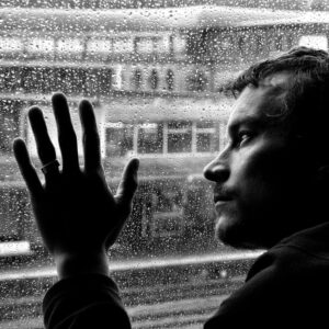 Un homme triste, joue appuyée sur une vitre remplie de goutelettes d'eau regarde le regard vide la pluie à l'extérieur