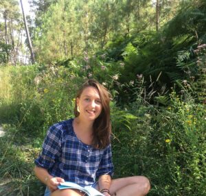 Photo de Julie, souriant, assise au sol et un livre posé sur ses genoux dans les bois