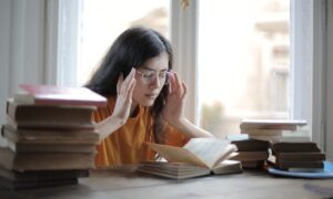 Une jeune femme assise à son bureau entre des piles de livres se tient les tempes, les yeux fermés, pour se concentrer